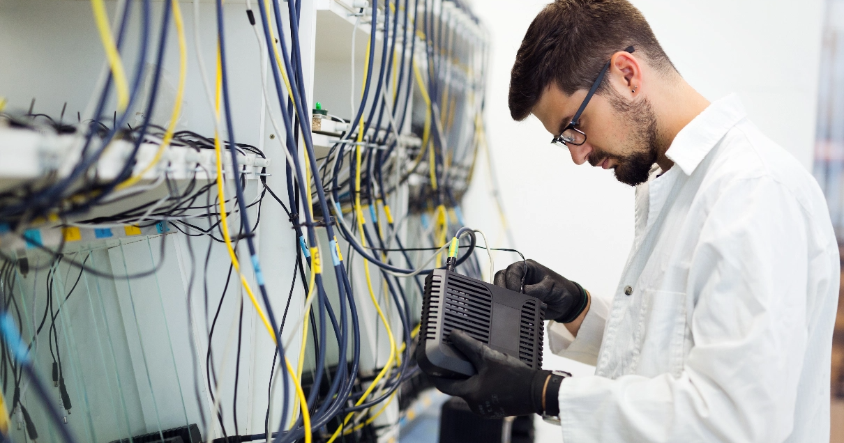 An IT tech wearing black gloves stands infront of a rack full of cables while plugging a cable into a modem.