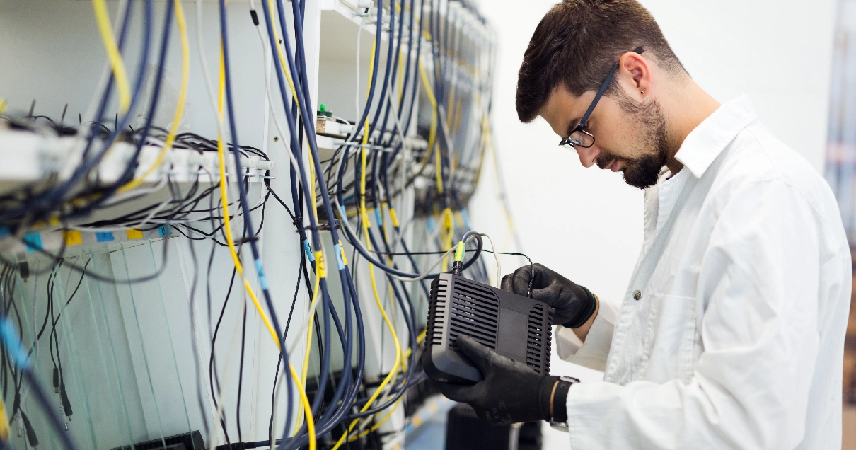 Un technicien informatique portant des gants noirs se tient devant un rack rempli de câbles et branche un câble sur un modem.