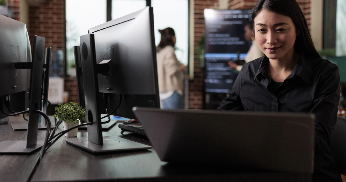 A female cybersecurity expert sits infront of a laptop and monitors in an open office.